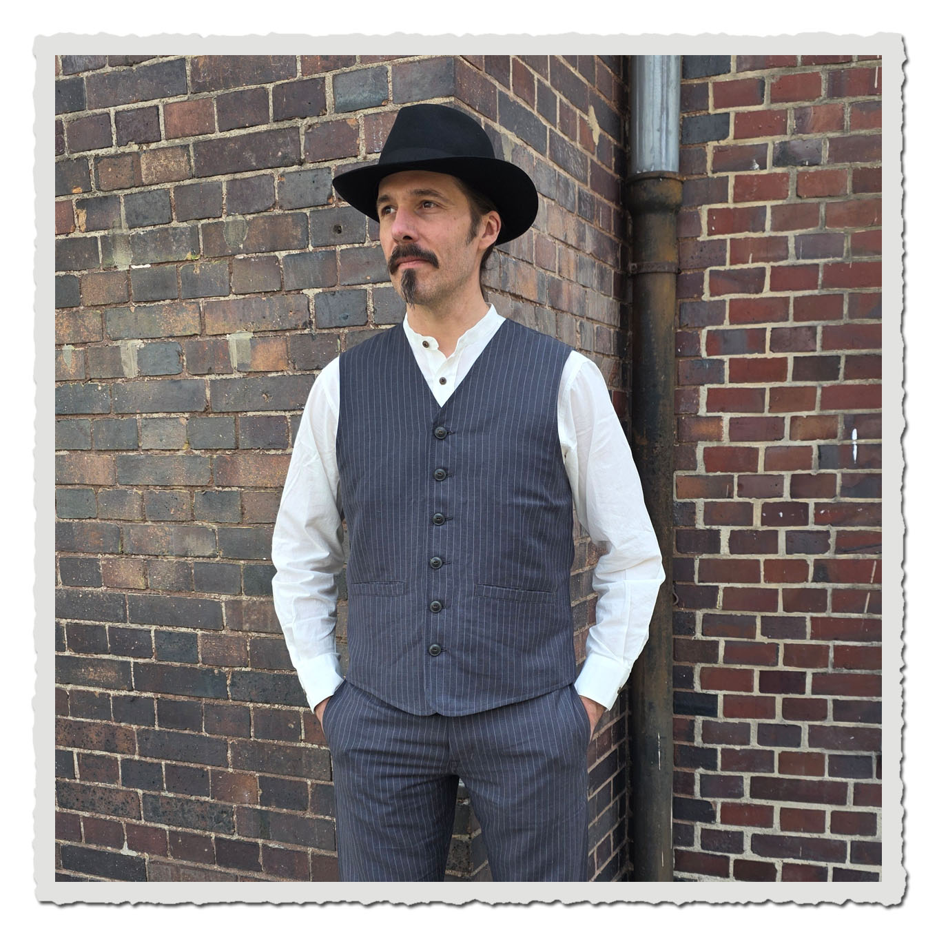 Model in grauer Nadelstreifenweste und weißem Hemd vor dunkler Backsteinwand im Außenbereich.